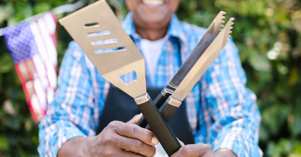 Elderly Man Holding Barbecue Utensils and Standing in the Garden Decorated for the 4th of July